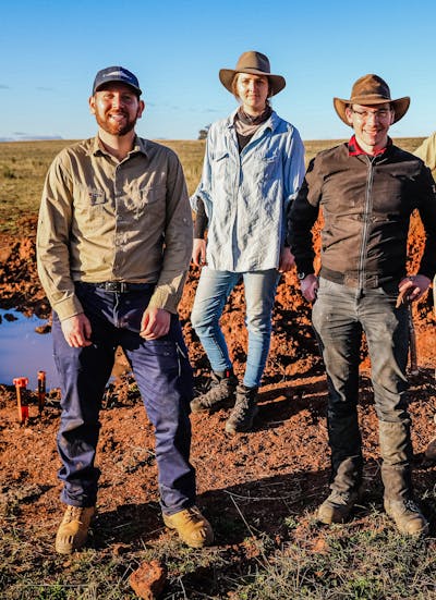 Three people, two men standing on either side of one woman, in a rural field wearing outdoor gear with work boots and wide brimmed hats.