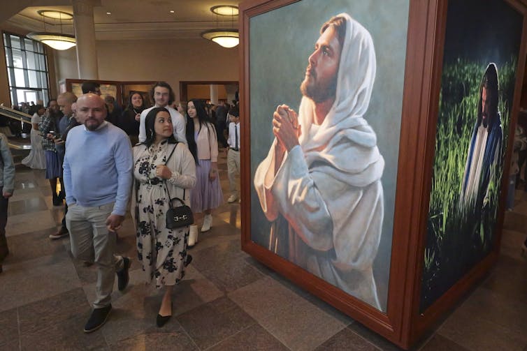 A couple dressed nicely and holding hands walks by a large portrait of Jesus, portrayed as a bearded white man, inside a large hallway.