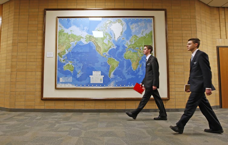 Two men in suits walk by a large map of the world framed on the wall of a hallway.