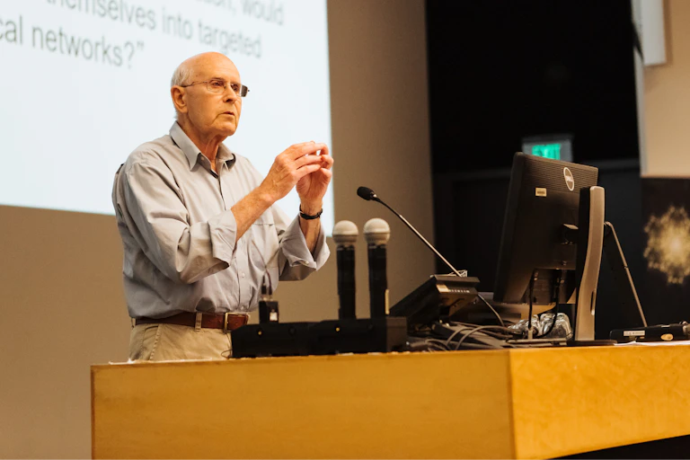 An older gentleman with glasses gives a talk in a lecture theatre.