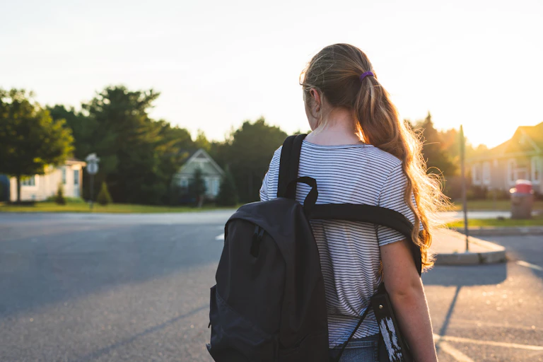 A young girl wears a backpack (dropping off one shoulder) in a carpark.