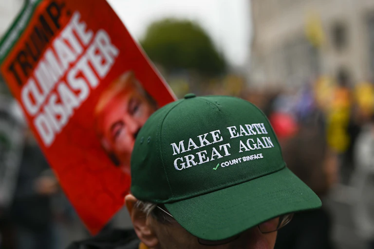 A person wearing a green cap that says 'Make earth great again' stands in front of an out-of-focus climate protest sign.