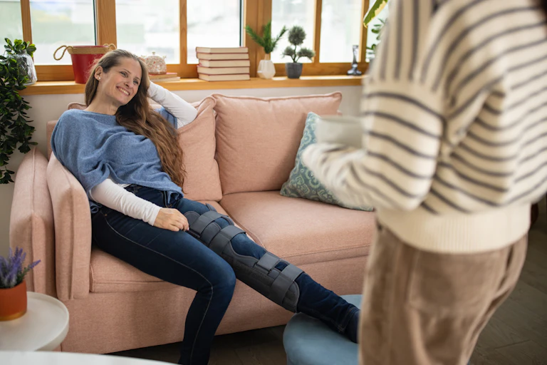 Woman brings a cup of tea to another woman on the couch with leg in a brace