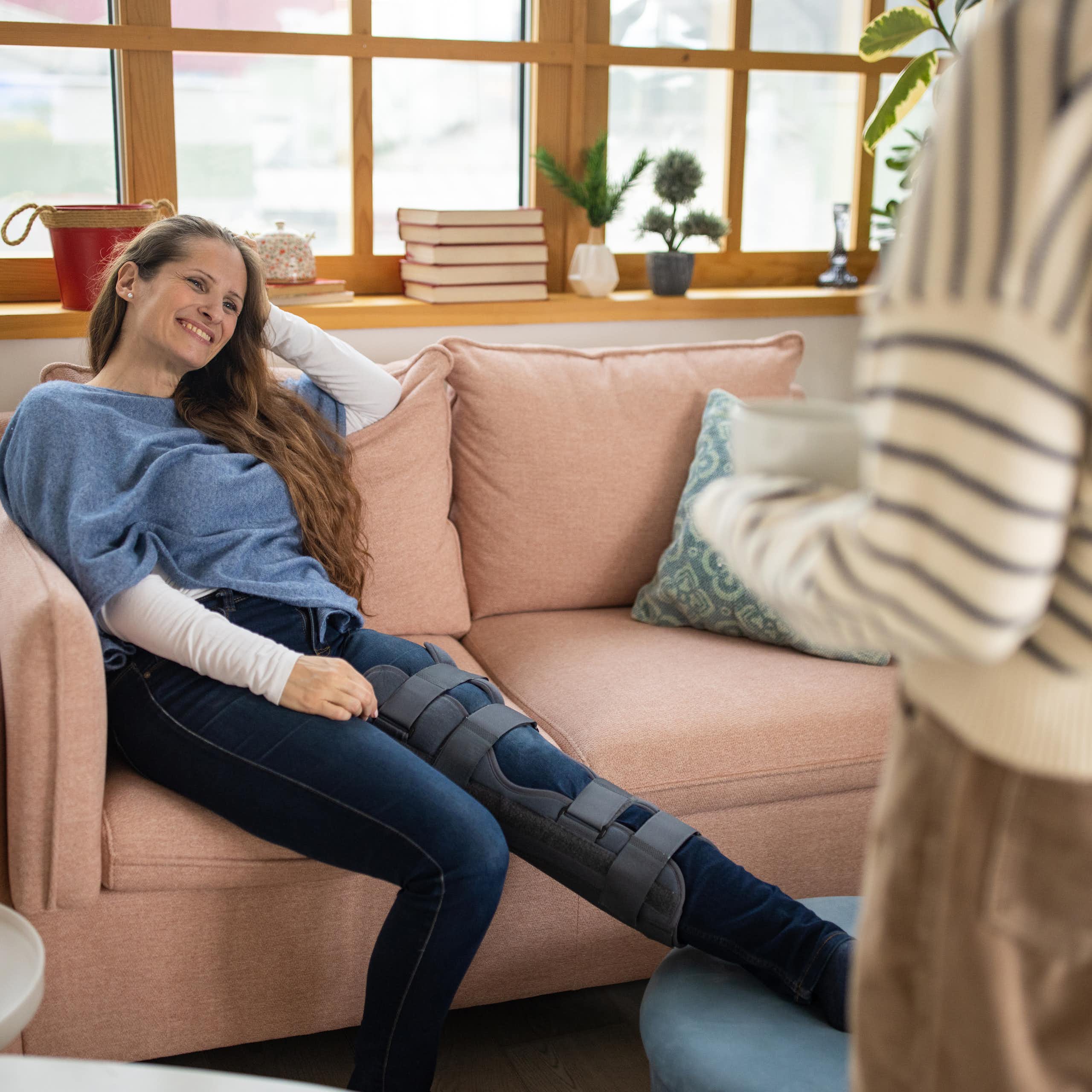 Woman brings a cup of tea to another woman on the couch with leg in a brace