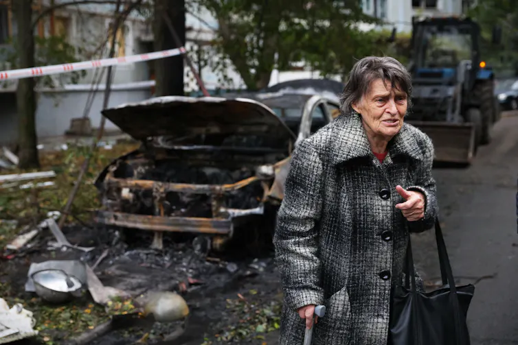 La mujer mayor con abrigos parece revelada junto al coche destruido.