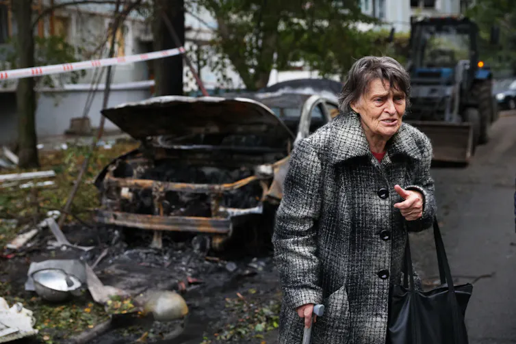 An older woman in an overcoat looks distraught next to a destroyed car.