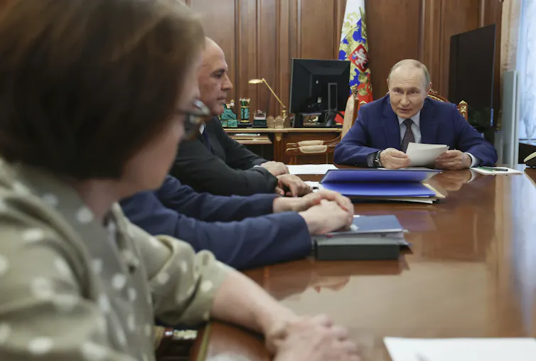 A balding man at the end of a wooden table peers at a woman whose back is to the camera.