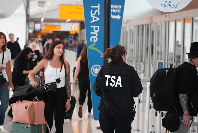 A person in a black jacket with TSA on the back guides travellers through an airport