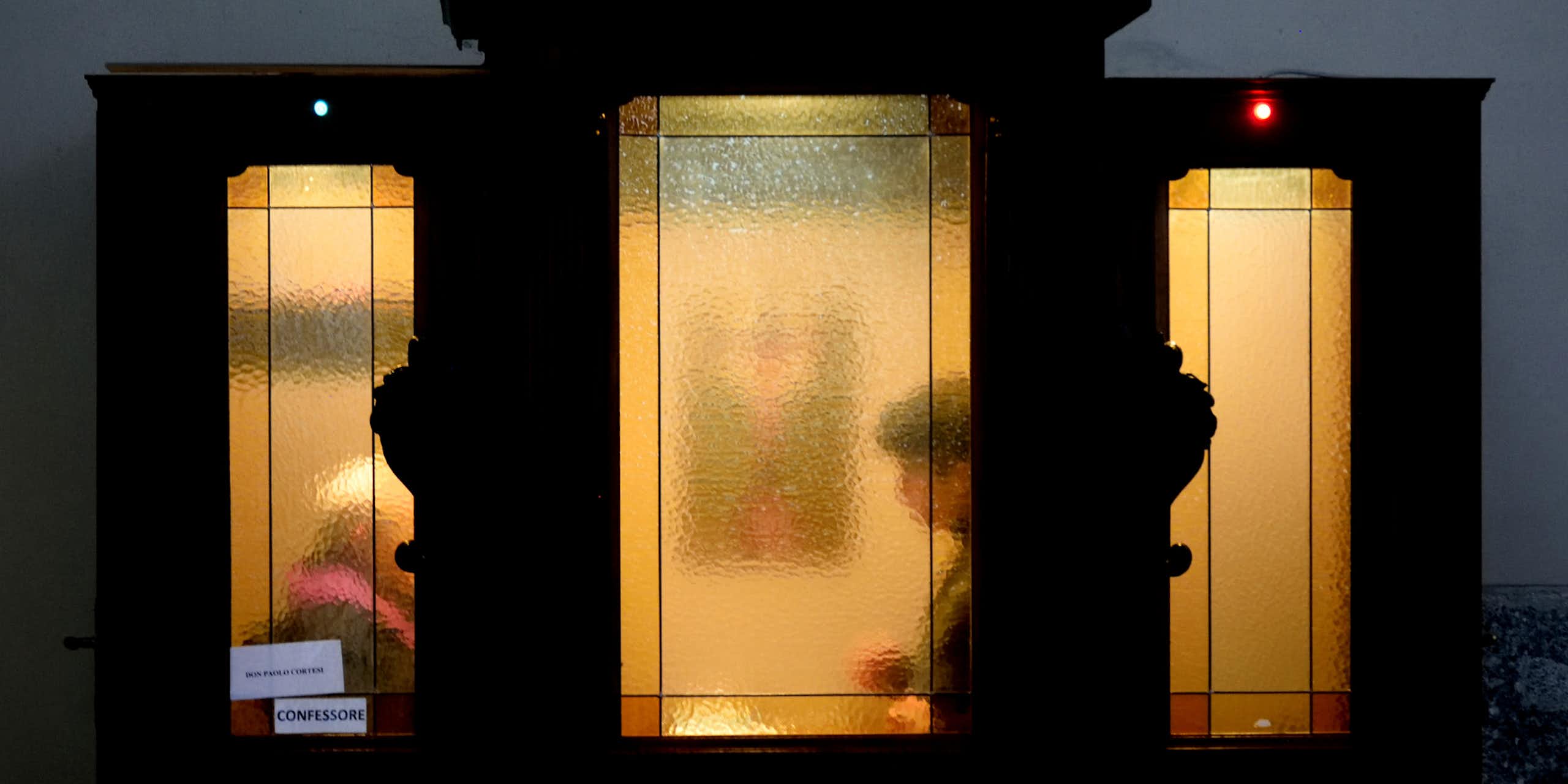 Two figures seated a few feet away from each other are seen through cloudy glass windows in an ornate wooden booth.
