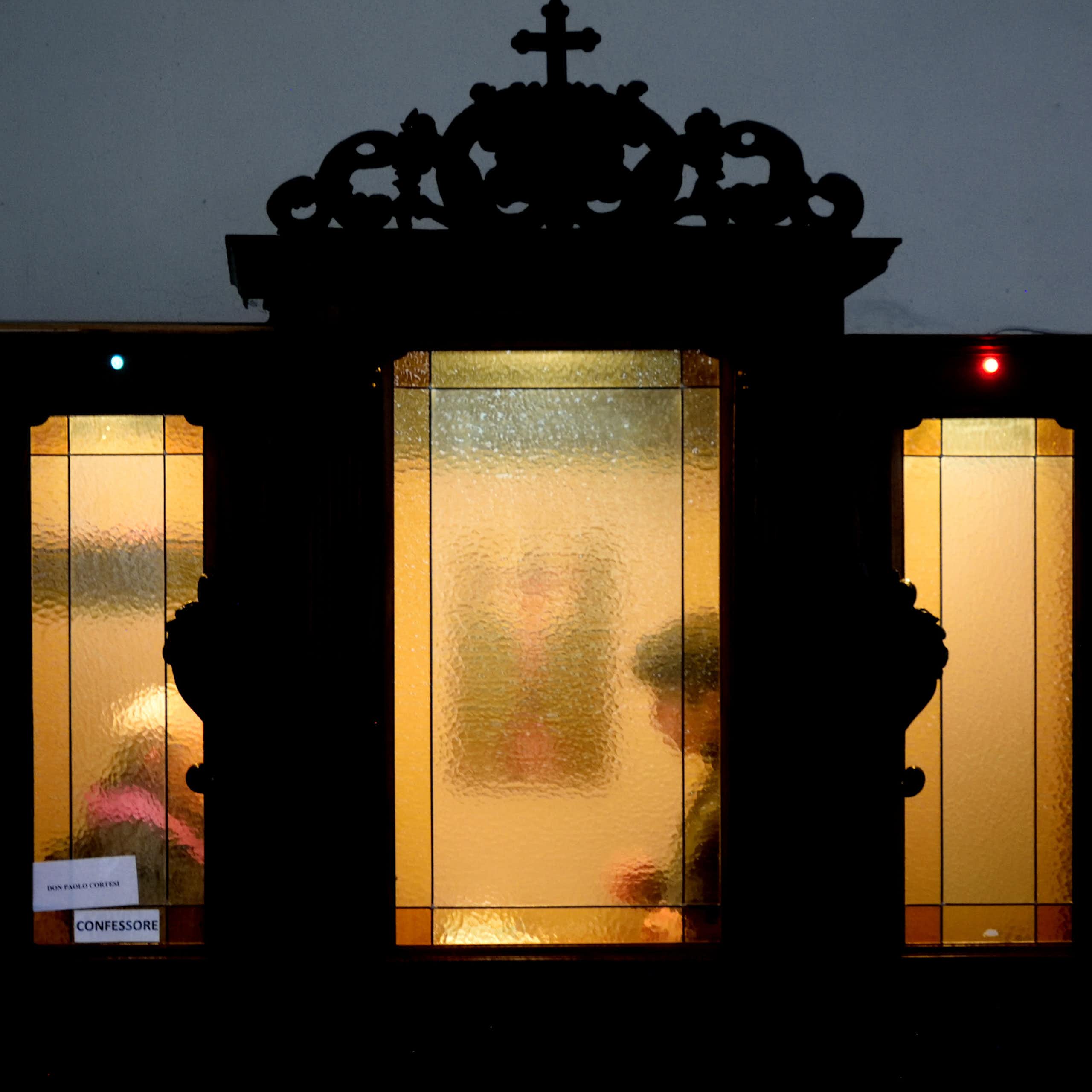 Two figures seated a few feet away from each other are seen through cloudy glass windows in an ornate wooden booth.