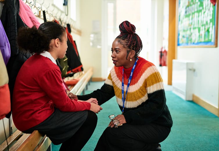 A teacher in a striped sweater crouches as she speaks with a girl, seated on a bench, who wears a red sweater and black skirt.