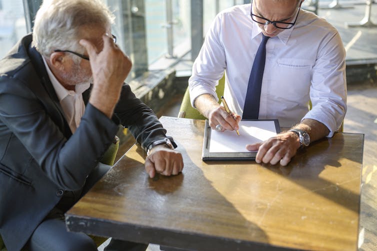 An older man in a suit holds his head in his hand as he sits at a table with a younger man taking notes.