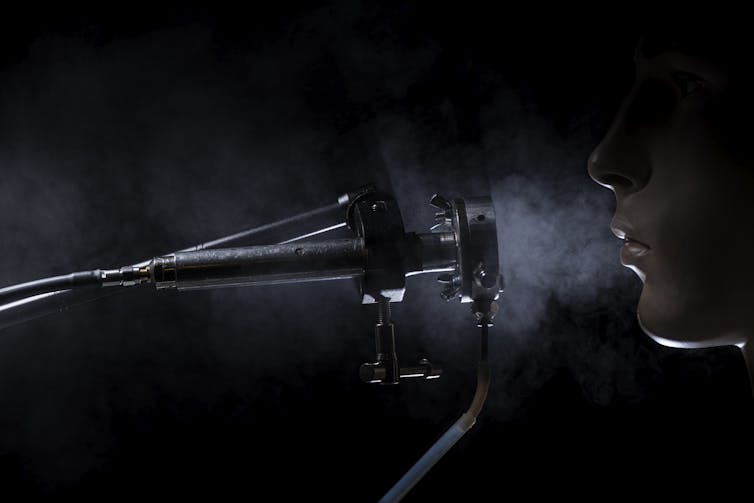 A photo showing a tube-like device, and a person exhaling into it out of their nose. A cloud of breath hangs in the air between.