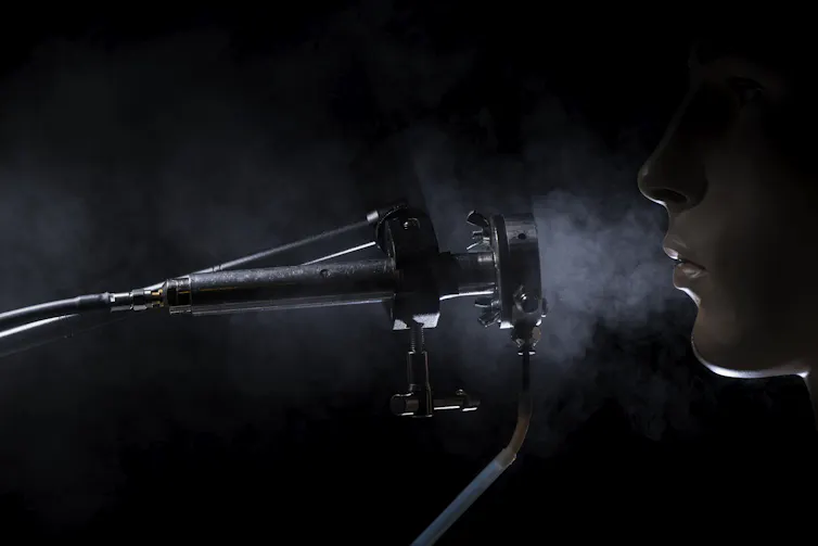 A photo showing a tube-like device, and a person exhaling into it out of their nose. A cloud of breath hangs in the air between.