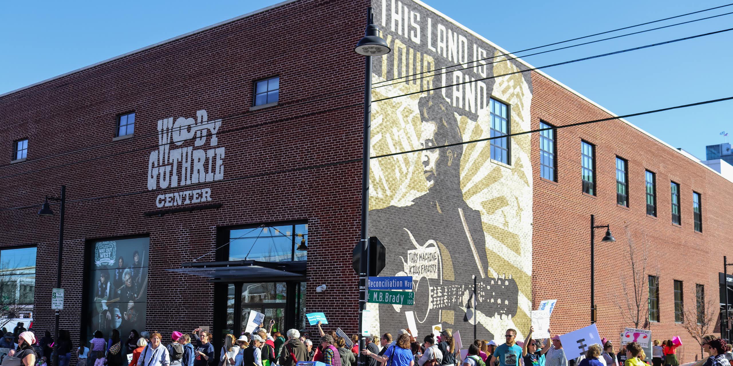 People hold signs and gather outside the Woody Guthrie Center, a brick building with a mural of the folksinger on one side, in daylight.