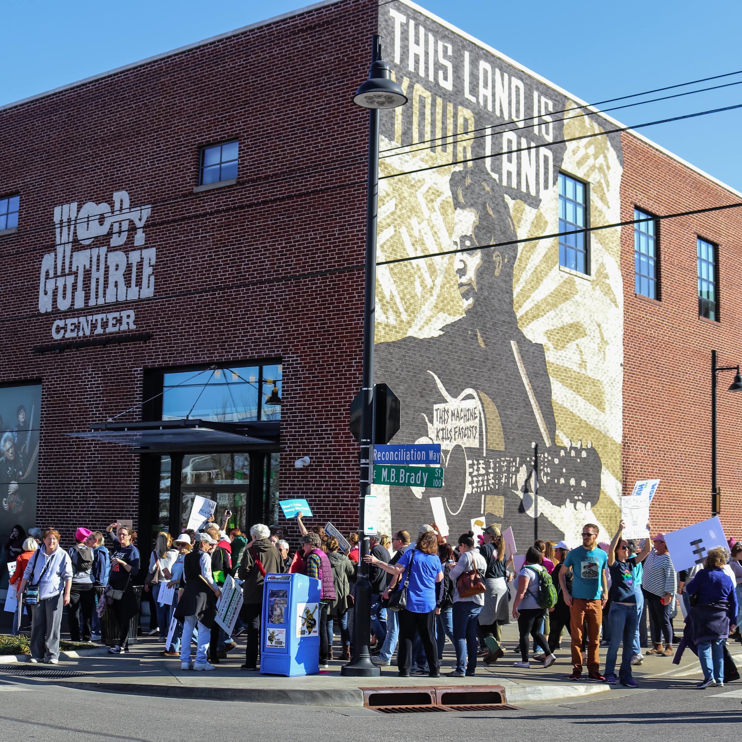 People hold signs and gather outside the Woody Guthrie Center, a brick building with a mural of the folksinger on one side, in daylight.