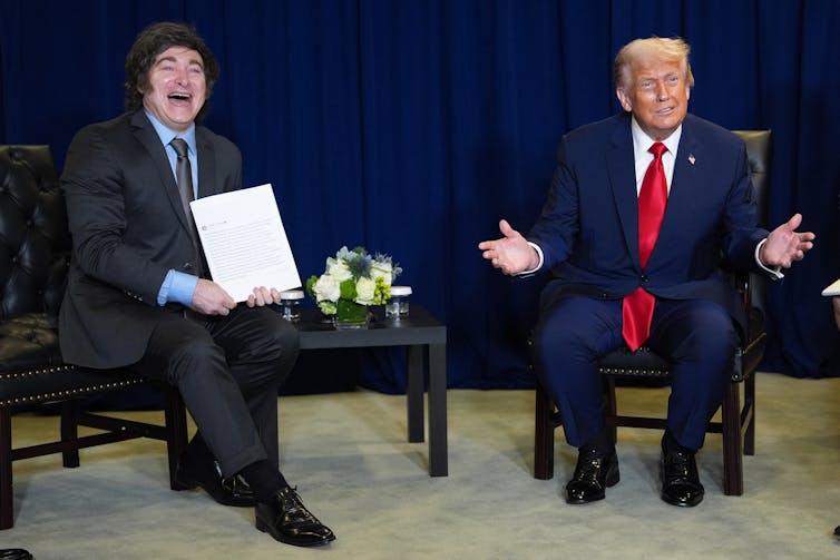 Milei and Trump seated on a stage. Milei is holding a piece of paper up.