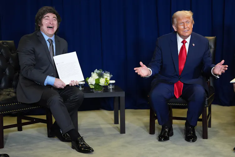 Milei and Trump seated on a stage. Milei is holding a piece of paper up.