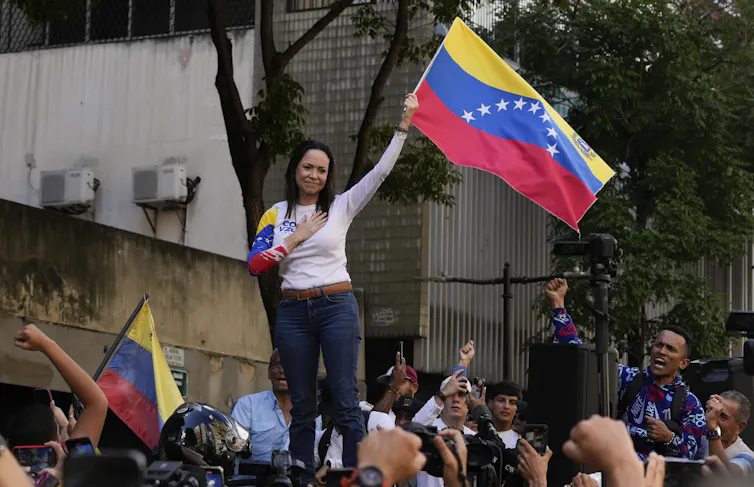 A dark-haired woman holds a yellow, blue and red flag smiling with a hand to her heart.
