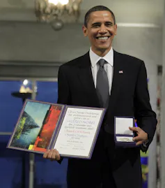 A Black man stands smiling and holding a medal and diploma.