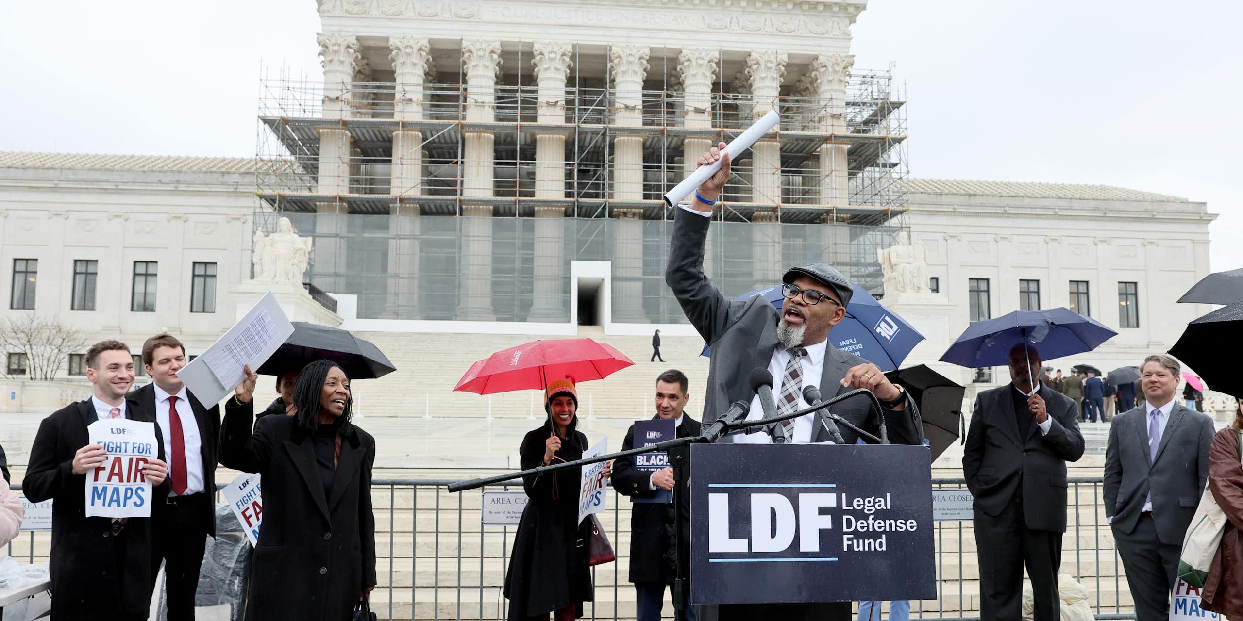 A group of people standing in the rain in front of a large, elegant building, as one of them speaks at a lectern.