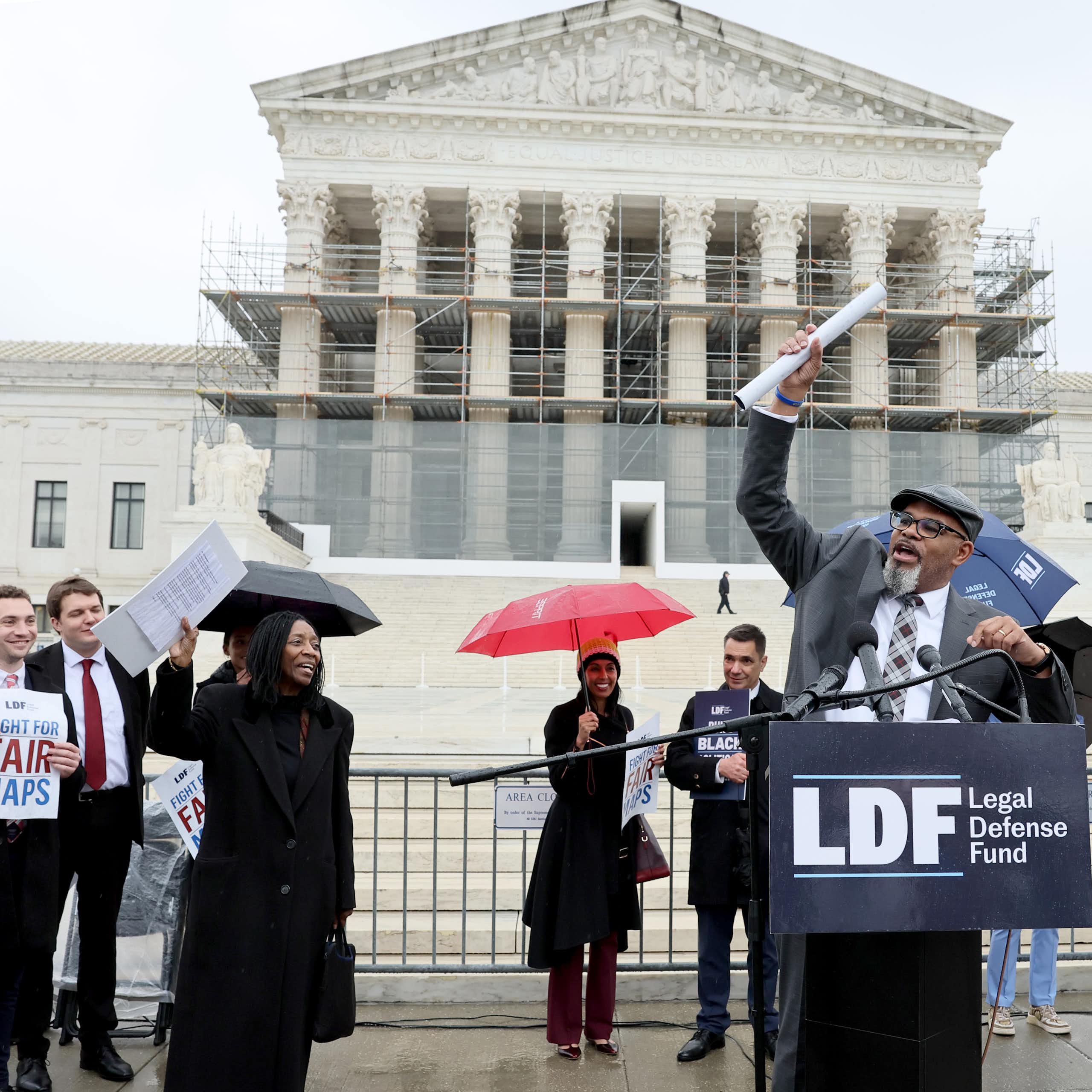 A group of people standing in the rain in front of a large, elegant building, as one of them speaks at a lectern.