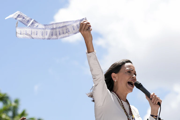 A dark-haired woman holds up pieces of paper while talking into a microphone.