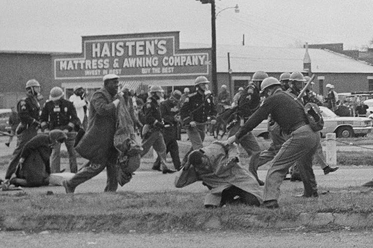 Police smashing marchers on a street with billy clubs.