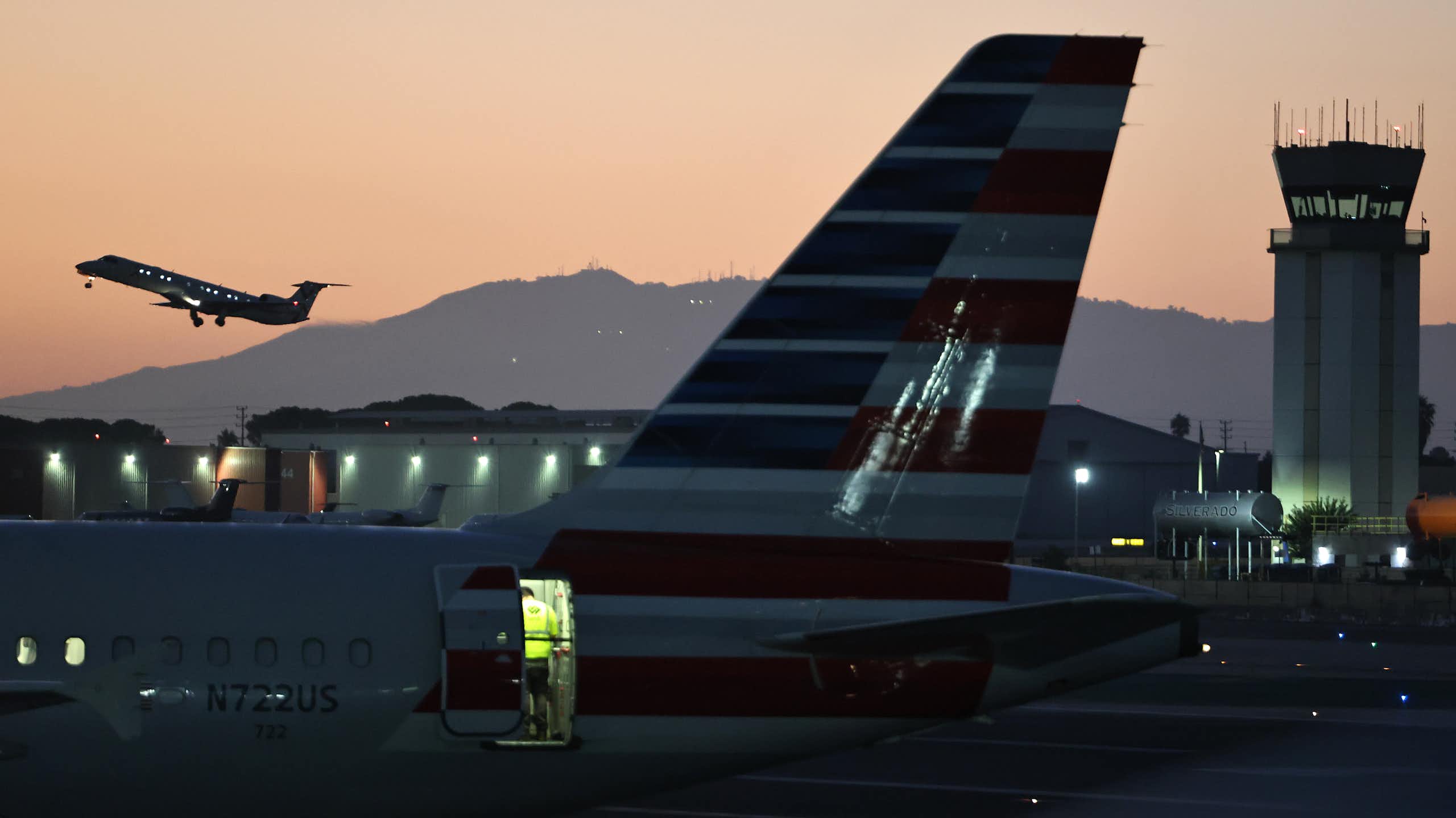 a plane is seen taking off in the distance while in the foreground are a plane on the tarmac and an air traffic control tower.