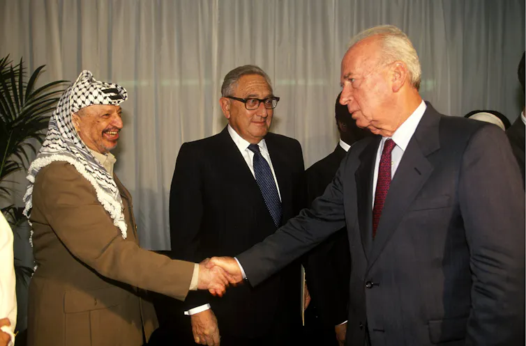Three men stand, two shaking hands.