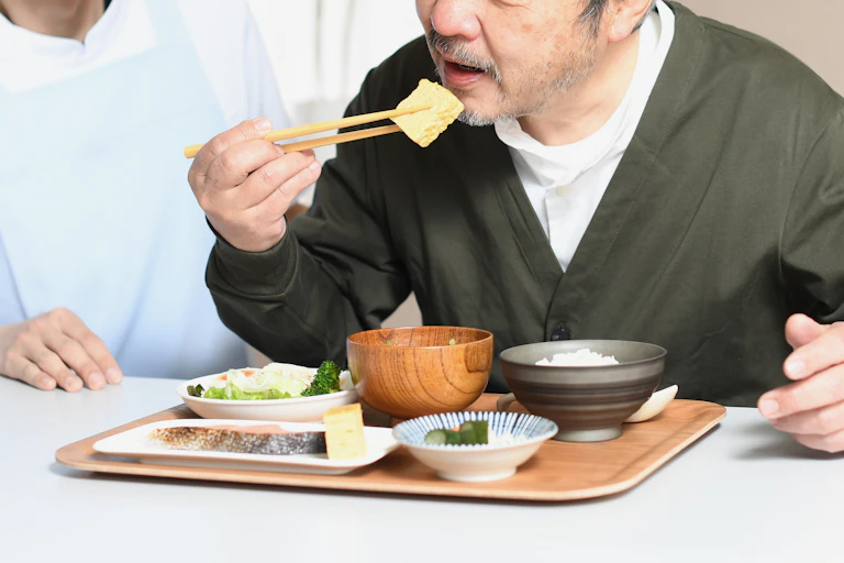 Older man holds a piece of tofu with chopsticks above a tray of small dishes.