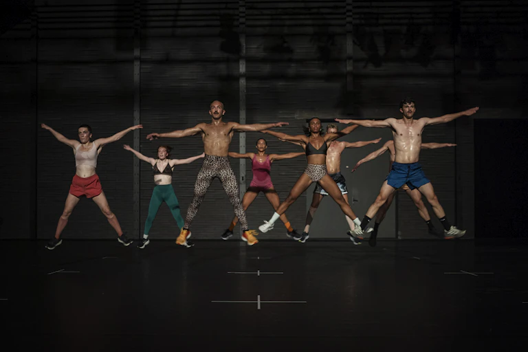 A group of dancers in exercise gear do star jumps.