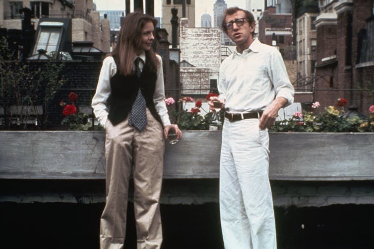Un hombre de pantalón y camisa blanca y una mujer de pantalón beis, chaleco y corbata, hablan en una terraza.