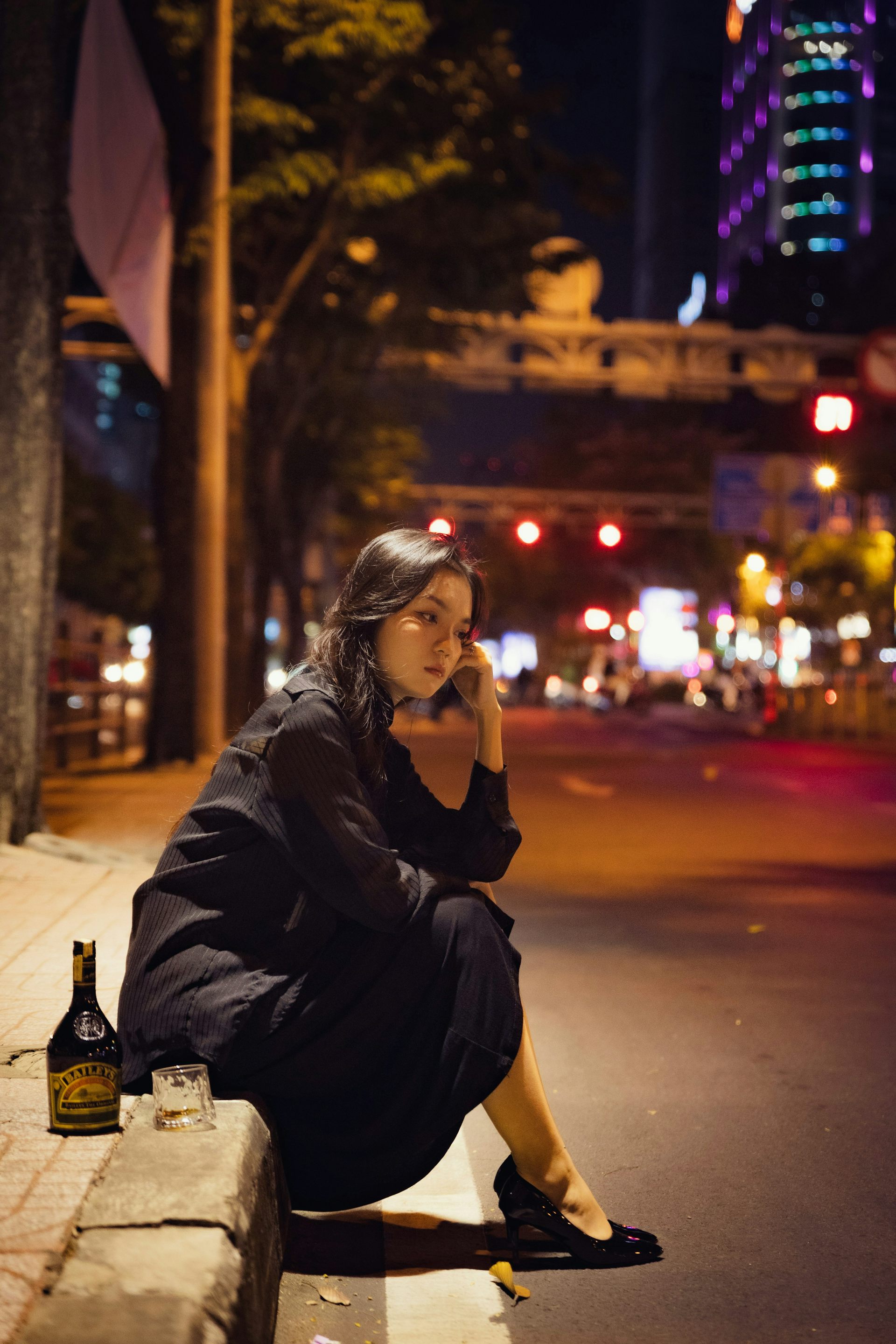 Photo d’une jeune femme à l’air pensif et triste assise au bord d’un trottoir avec une bouteille et un verre posés à côté d’elle