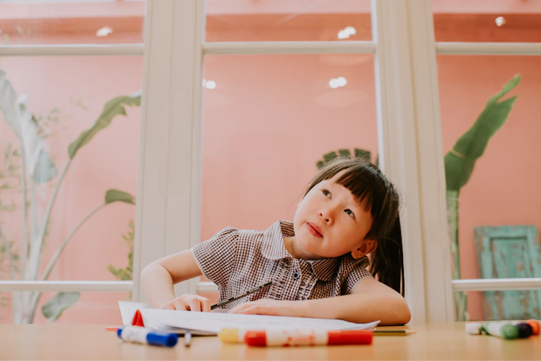 Young child sitting at a table and looking up toward the sky as if daydreaming.