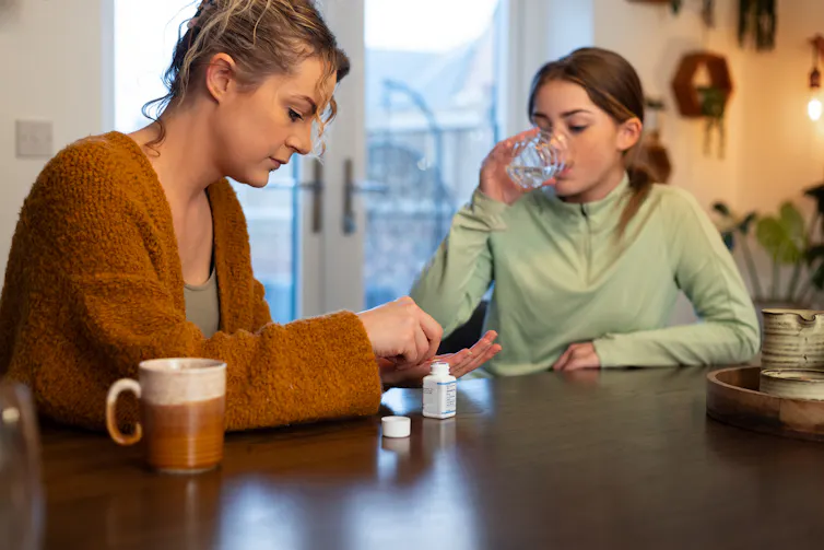 Middle-aged woman sitting at a table and giving a pill to an adolescent girl, who is sipping a glass of water.