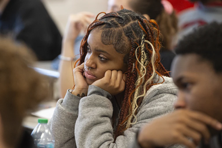 High school student sits at a desk in a classroom with her hands propping up her face.