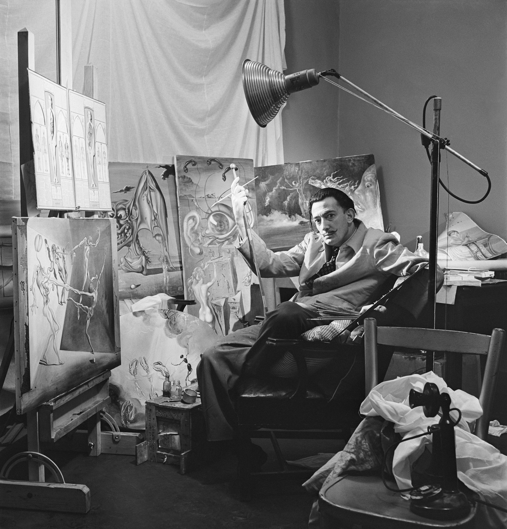 Black and white photo of man in suit sitting in chair in front of paintings on canvases