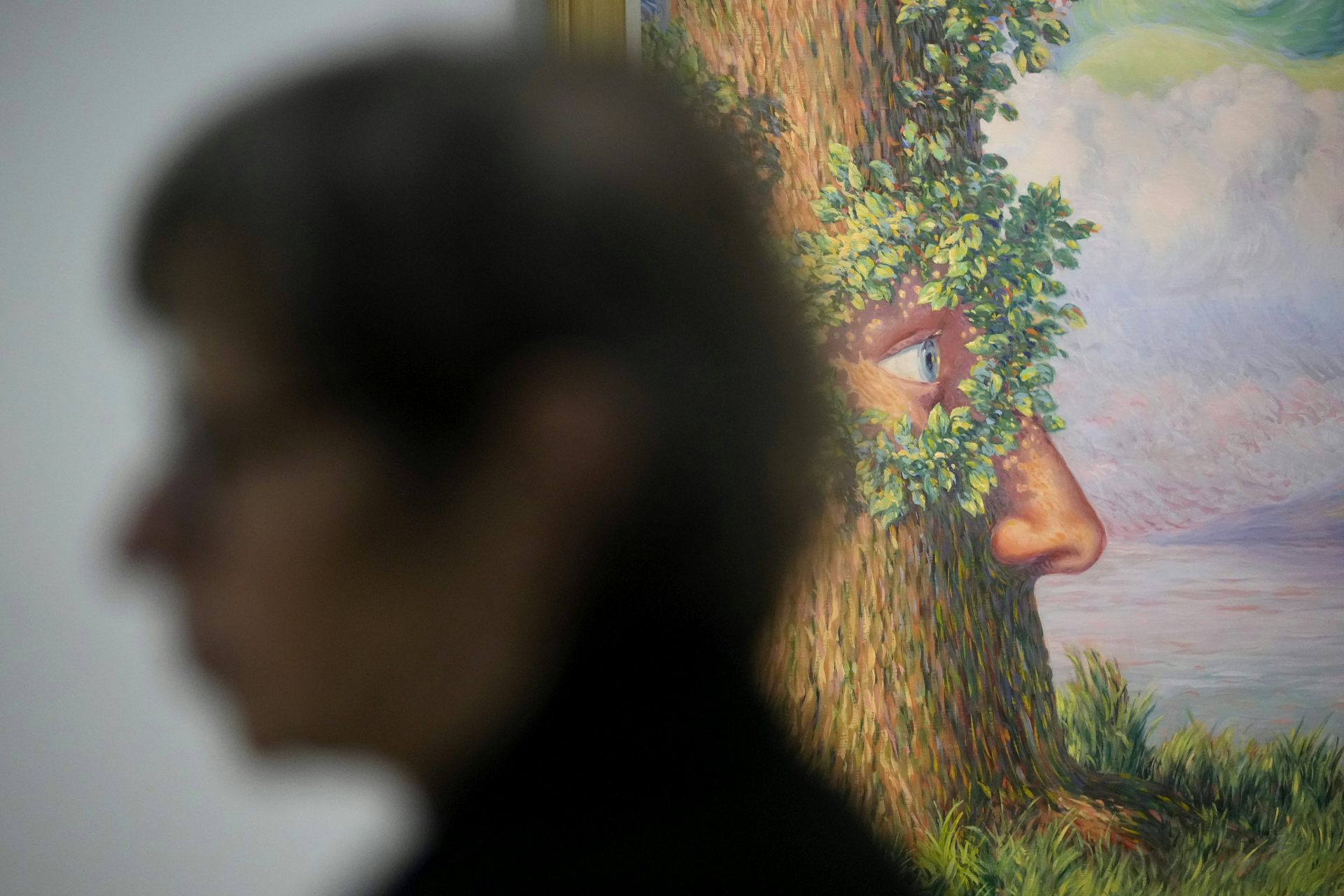 Woman in foreground, blurred, shown passing a colorful painting of tree with human face