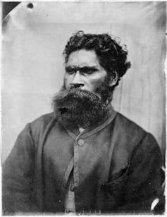 An old black-and-white portrait photograph shows Aboriginal artist William Barak posing. He has a large beard and moustache.