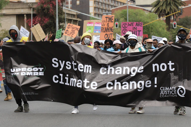 A group of a few hundred people march down a city street, carrying a banner that says 'System change not climate change'