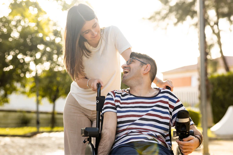 Un jeune homme est assis dans un fauteuil roulant. À ses côtés, se trouve une jeune femme. Les deux jeunes sont dans un parc au coucher du soleil et ils se regardent joyeusement.