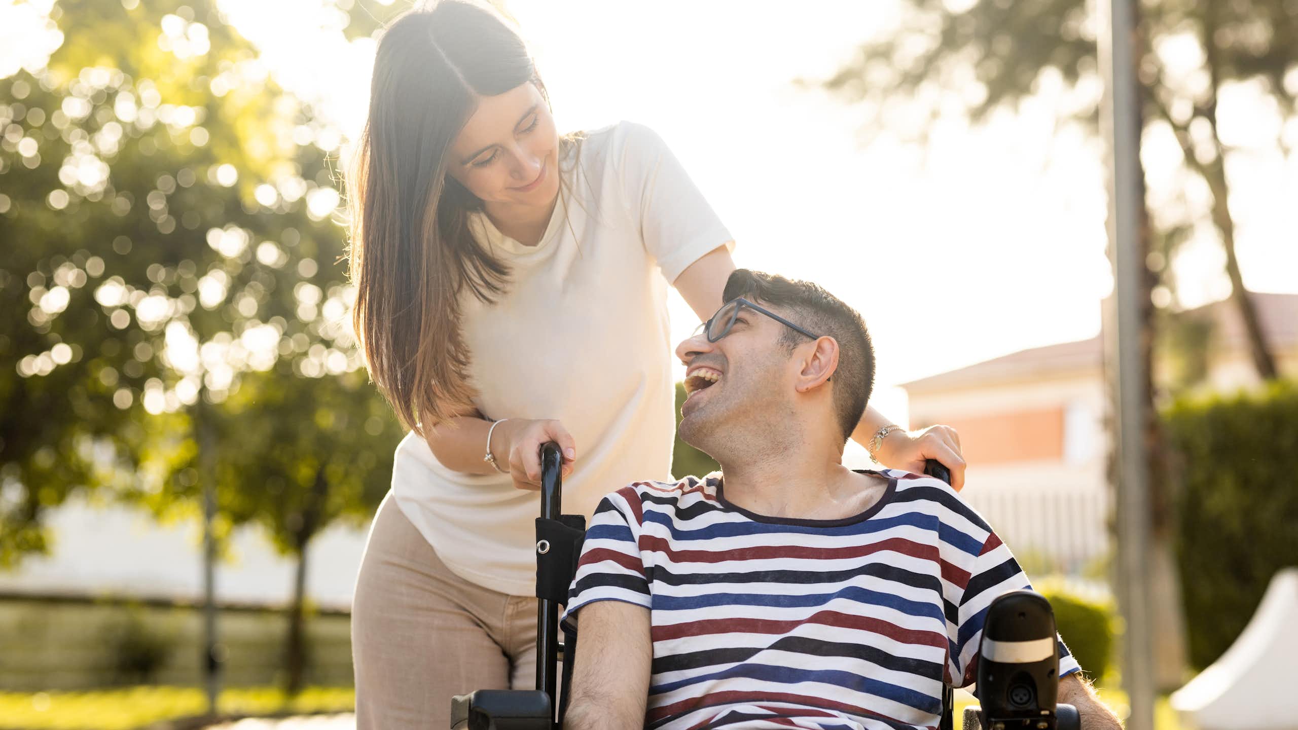 Un jeune homme est assis dans un fauteuil roulant. À ses côtés, se trouve une jeune femme. Les deux jeunes sont dans un parc au coucher du soleil et ils se regardent joyeusement.