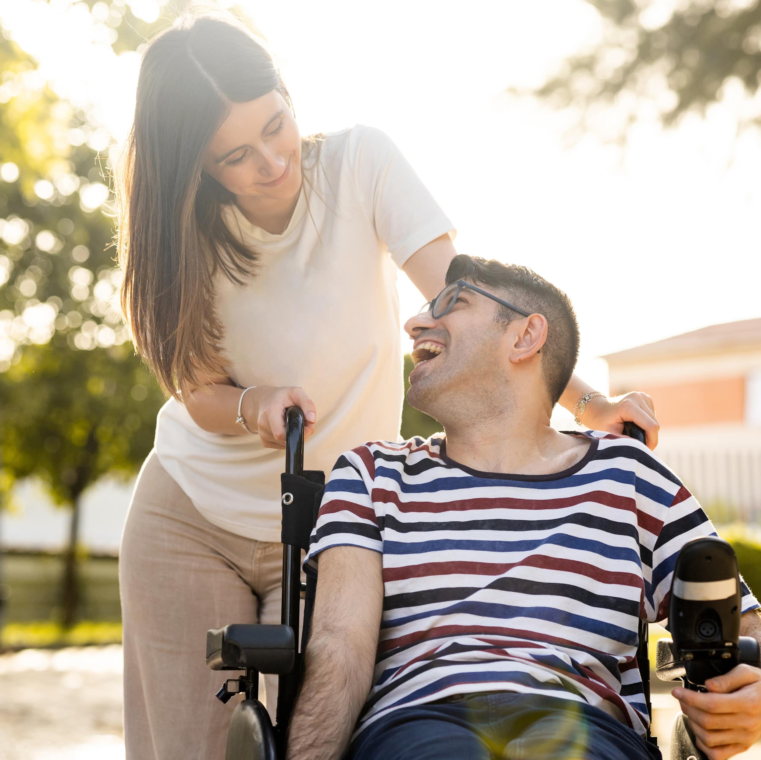Un jeune homme est assis dans un fauteuil roulant. À ses côtés, se trouve une jeune femme. Les deux jeunes sont dans un parc au coucher du soleil et ils se regardent joyeusement.