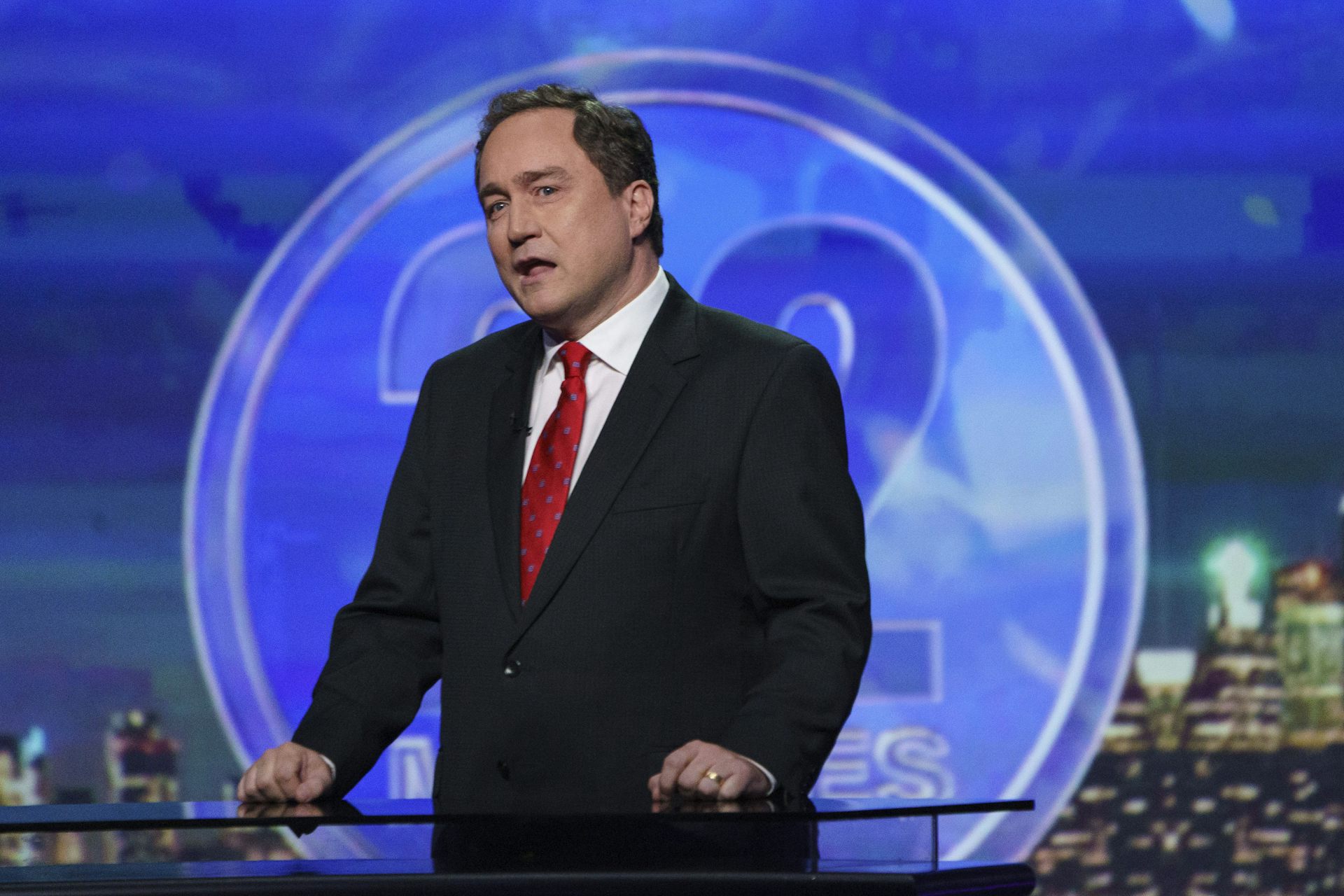 A man in a suit and red tie with a twinkle in his eye looks like he's ready to laugh against a blue background.