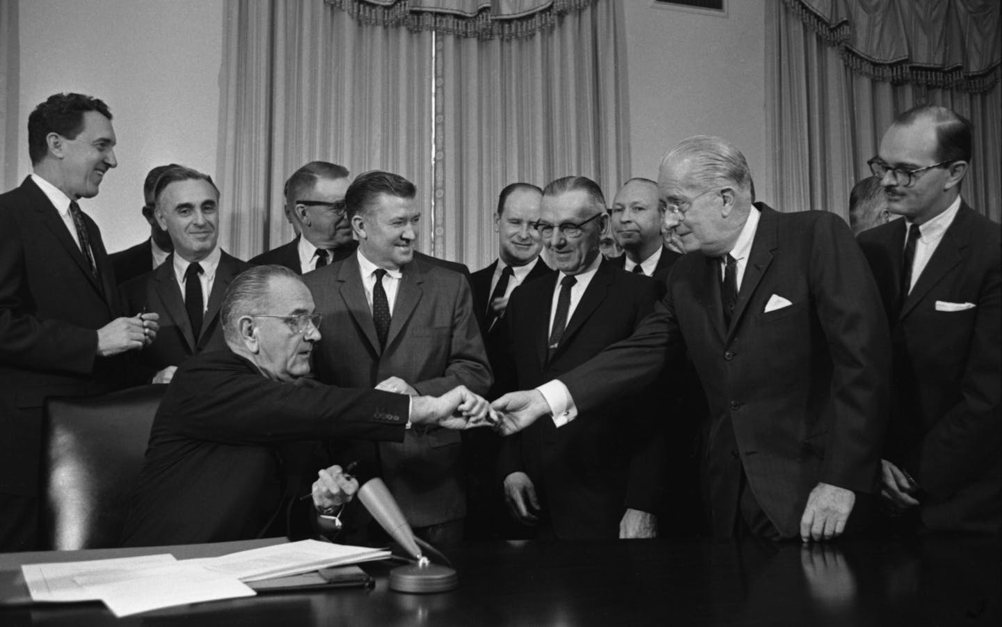 A group of men in suits stand around a seated man, who is handing a pen to one of the standing men.