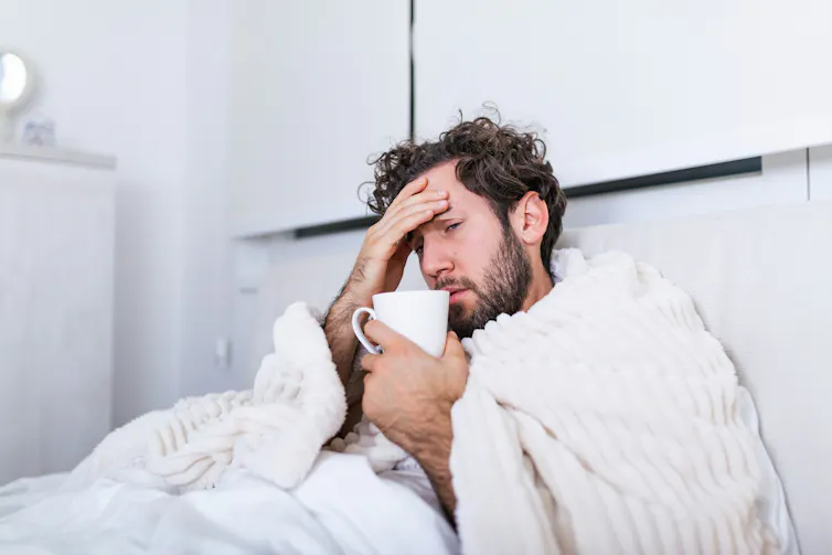 A man with a blanket wrapped around his shoulders sits in bed while nursing a cup of tea
