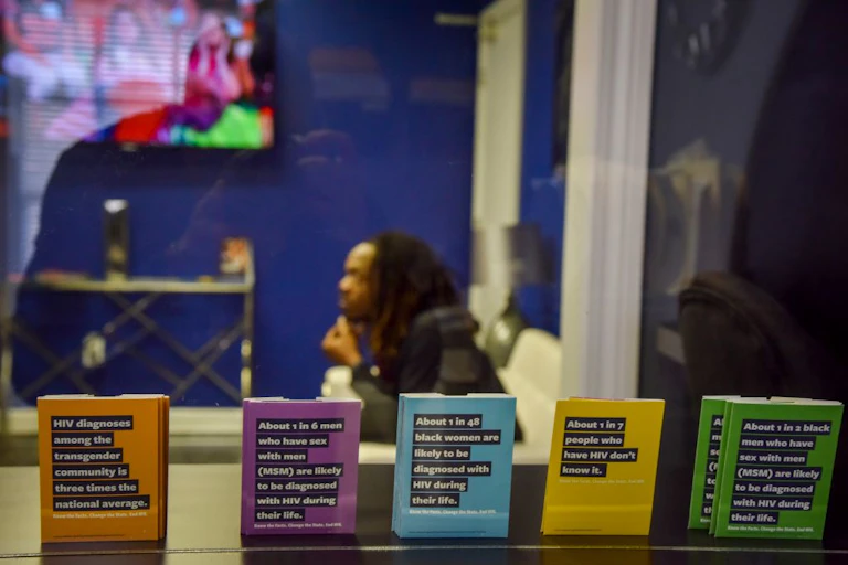 A Black man sitting behind the glass of a clinic waiting room, where displayed are multicolored pamphlets on HIV diagnosis statistics