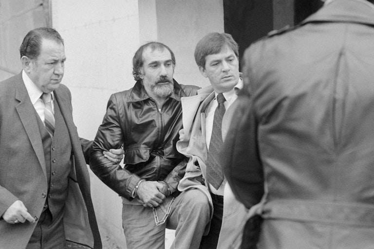 Black and white photo of men escorting handcuffed man with mustache and beard and wearing a leather jacket