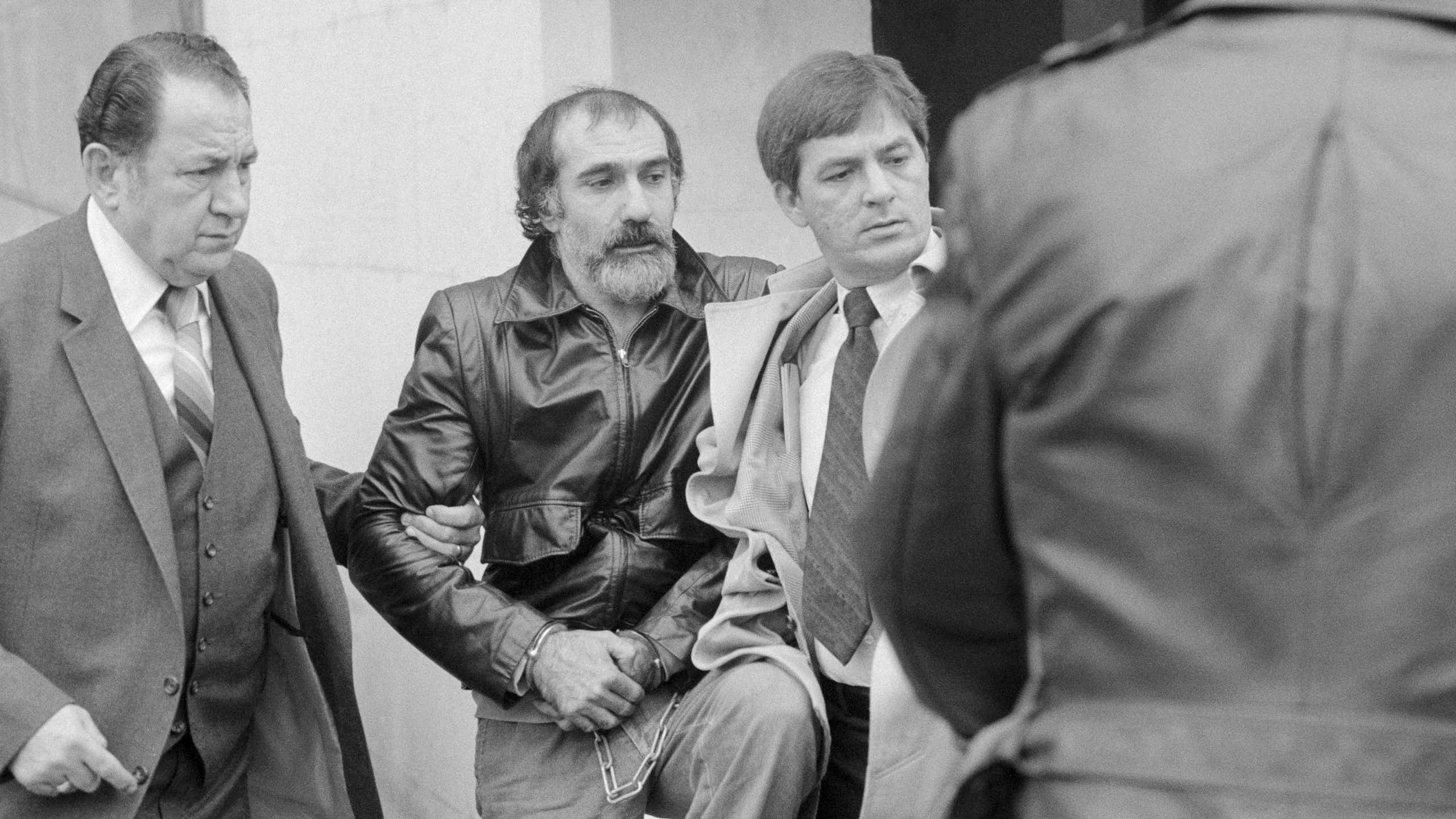 Black and white photo of men escorting handcuffed man with mustache and beard and wearing a leather jacket